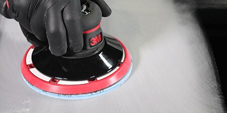 Una persona con guantes negros utiliza una lijadora eléctrica de marca 3M con un disco abrasivo azul para alisar una superficie y lograr un acabado pulido.
