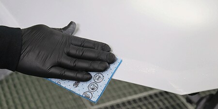 Una persona que lleva un guante negro utiliza una almohadilla abrasiva azul para lijar el borde de una superficie lisa y blanca.
