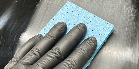 Una persona con guantes negros sostiene una almohadilla abrasiva azul y lija una superficie oscura para crear un acabado liso y uniforme.

