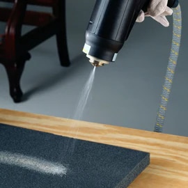 Image of a gloved hand spraying adhesive on a piece of black foam with a high-back chair frame in the background