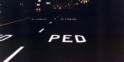 White illuminated pavement markings on dark road at night, reading “PED XING.”

