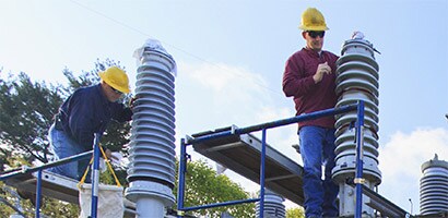 Dos hombres con cascos trabajan muy por encima del suelo en empalmes y terminales de alta tensión.

