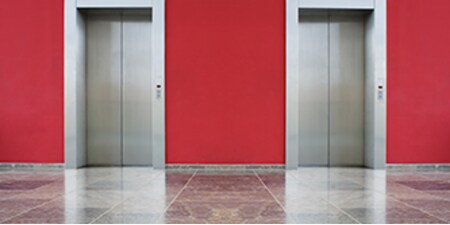 Imagen de las puertas de dos ascensores con puertas cerradas. Paredes rojas y piso gris brillante.
