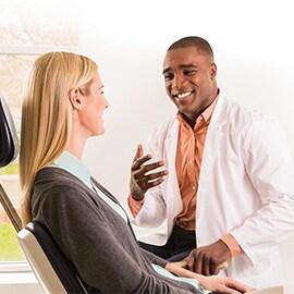 Black male doctor speaking with a blonde, white, female patient in the dentist chair.