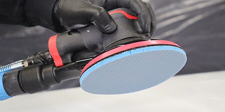Una persona con guantes negros sostiene una lijadora eléctrica equipada con un Disco Abrasivo Flexible de Espuma 3M™ Hookit™ azul, lista para usarla en una superficie.
