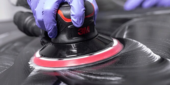 Una persona con guantes morados utiliza una lijadora eléctrica de marca 3M sobre una superficie oscura y crea un patrón circular.
